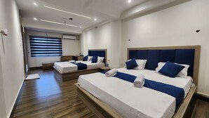 A hotel room with two beds, wooden flooring, a ceiling fan, and a window with blinds. - Lakhdatar Hotel (Deoghar)