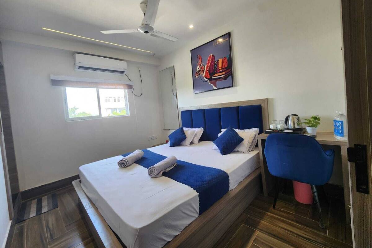 A hotel room with a bed, a desk, a chair, a ceiling fan, an air conditioning unit, a window, and a poster on the wall.