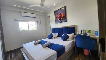 Habitación de hotel con una cama, un escritorio, una silla, un ventilador de techo, un equipo de aire acondicionado, una ventana y un cartel en la pared.