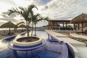 Ein Pool mit kreisförmigem Design, Palmen und strohgedeckten Sonnenschirmen.