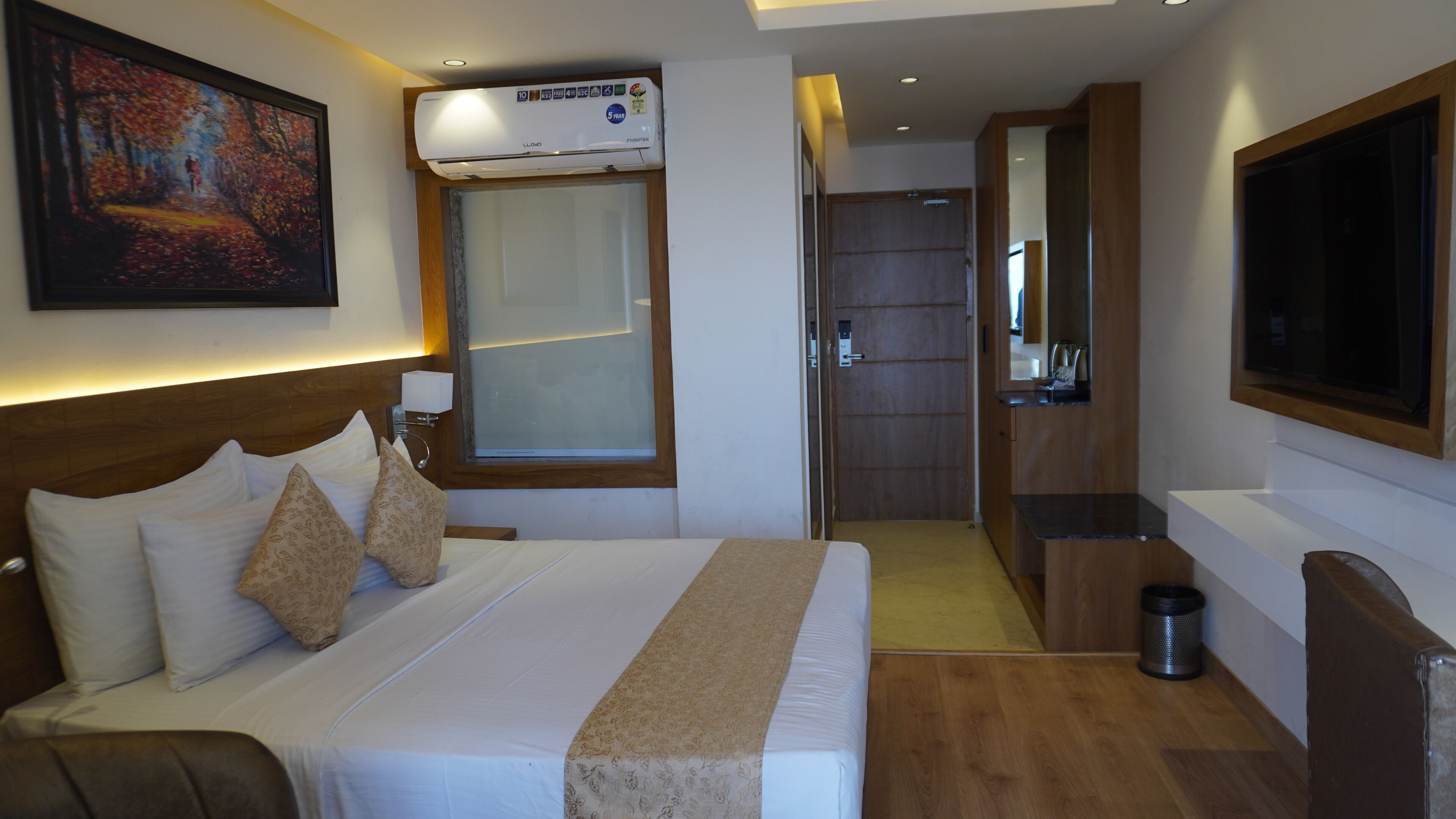 A hotel room with a large bed, a painting on the wall, an air conditioning unit, a TV, and a wooden floor.