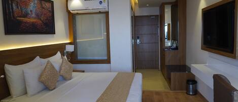 A hotel room with a large bed, a painting on the wall, an air conditioning unit, a TV, and a wooden floor.