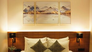A hotel room with a wooden headboard, two bedside lamps, a bed with white linens and leopard print pillows, and three framed paintings above the bed. - The novello by hotel evergreen  (Mussoorie)