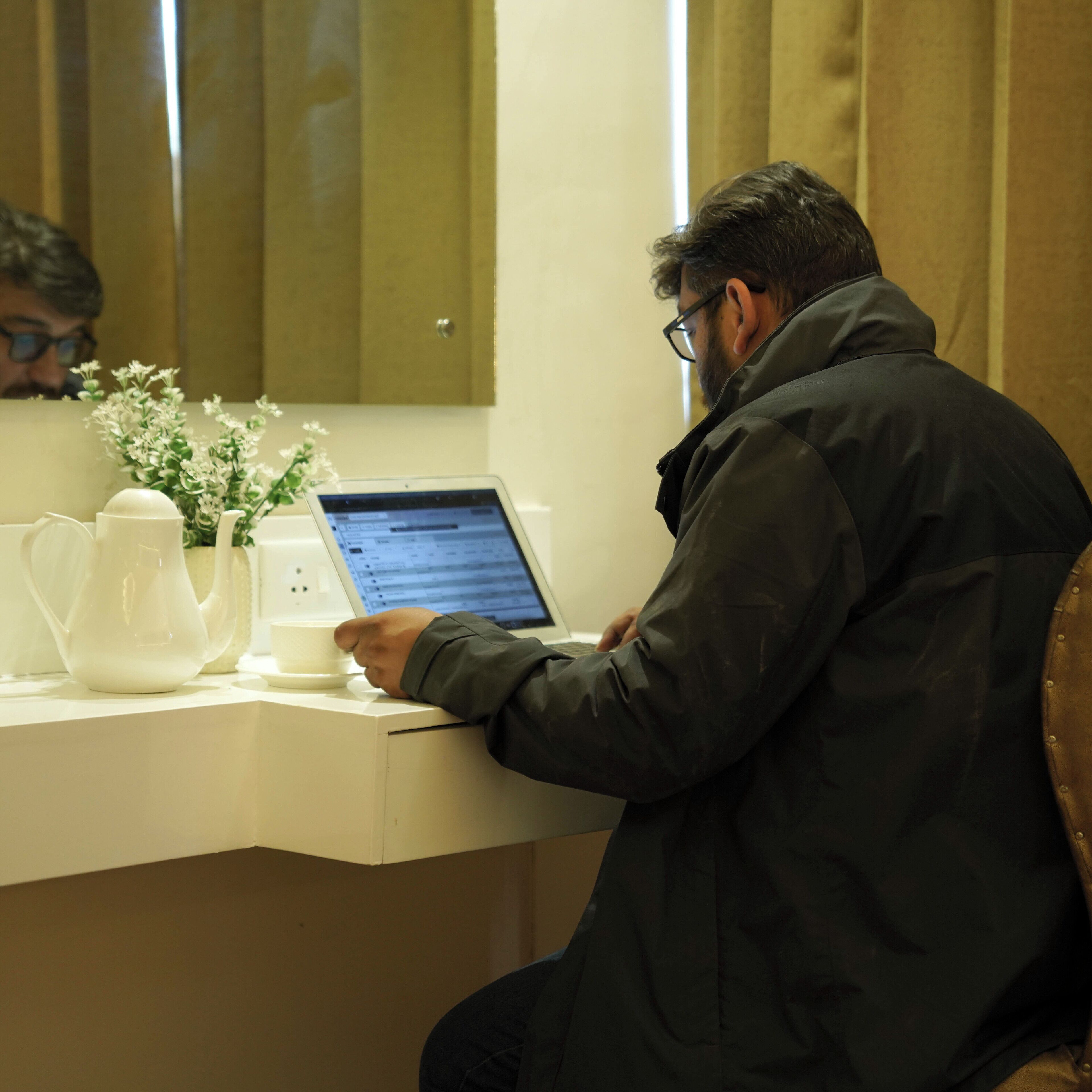 A person sitting at a desk with a laptop, a white teapot, and a small white flower arrangement.
