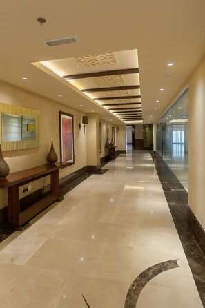 Un corridoio d'hotel con pavimento in marmo lucido, illuminazione incassata, una reception e un ascensore in vetro.
