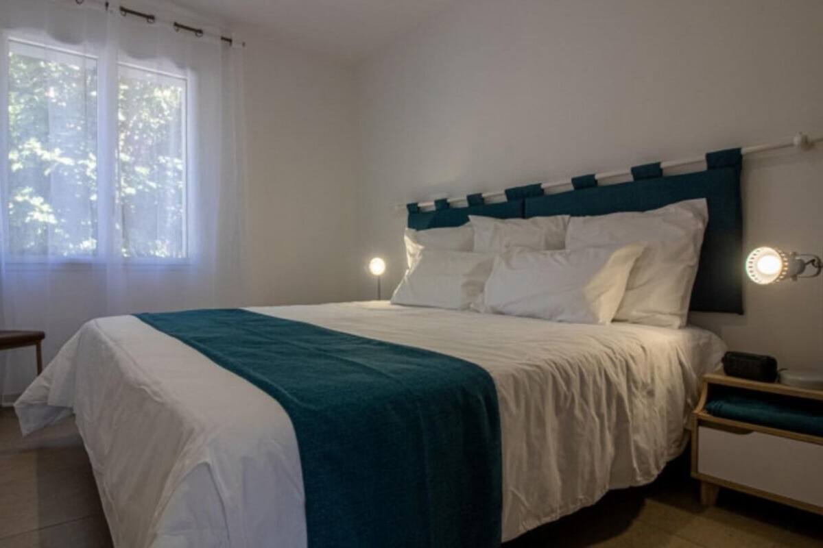 Un lit bien fait, avec une literie blanche et turquoise, une table de chevet avec une lampe et une chaise dans une pièce dotée d’une grande fenêtre.