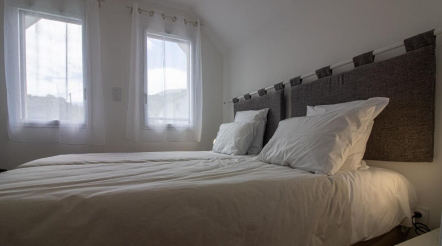 A bedroom with a large bed, two windows with sheer curtains, and a plain headboard.