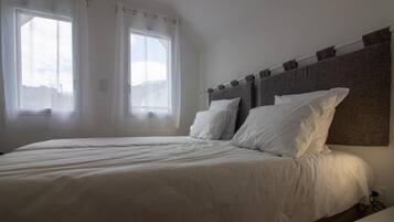 A bedroom with a large bed, two windows with sheer curtains, and a plain headboard.