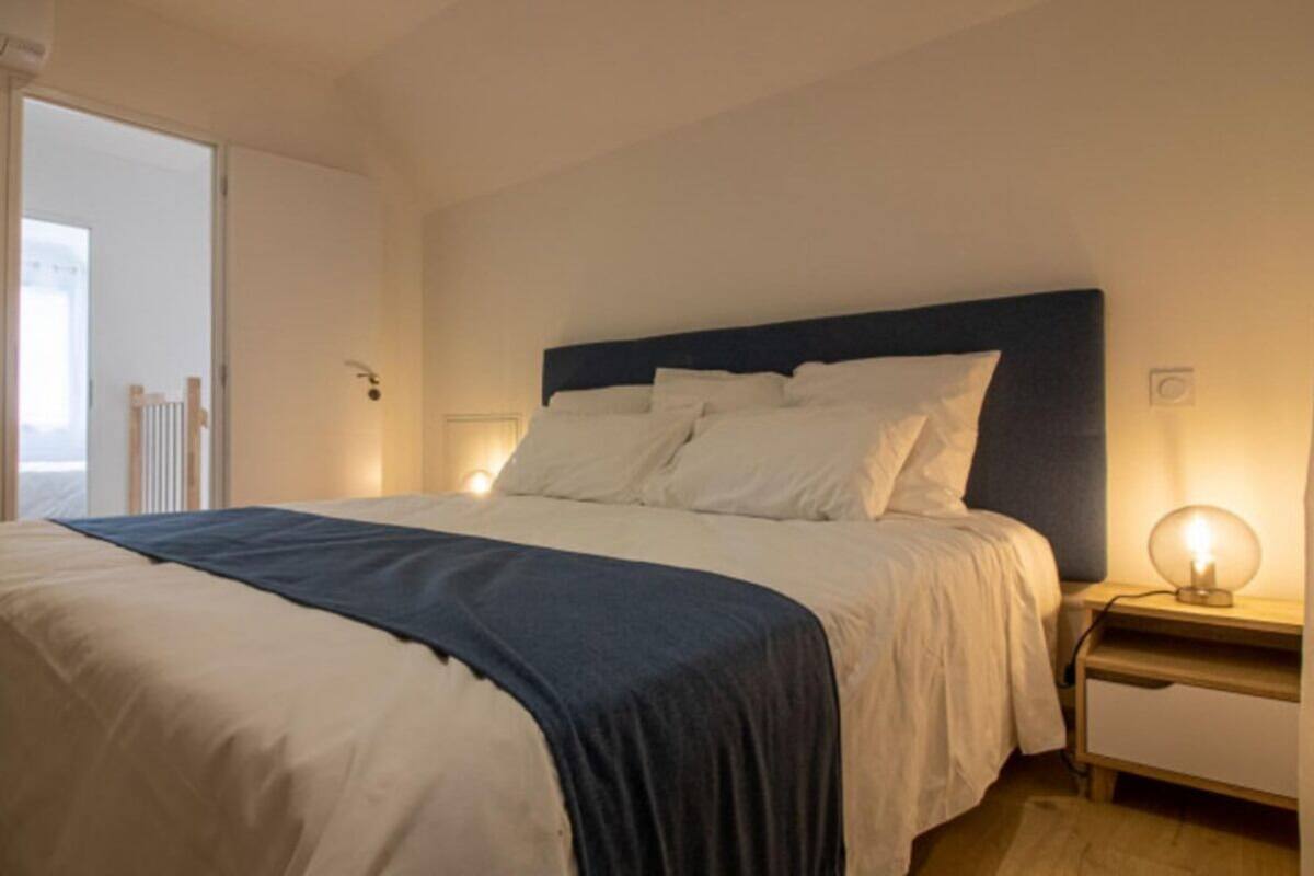 Un lit bien fait, recouvert d’une couverture bleue, une table de chevet avec une lampe, et une vue sur un balcon.