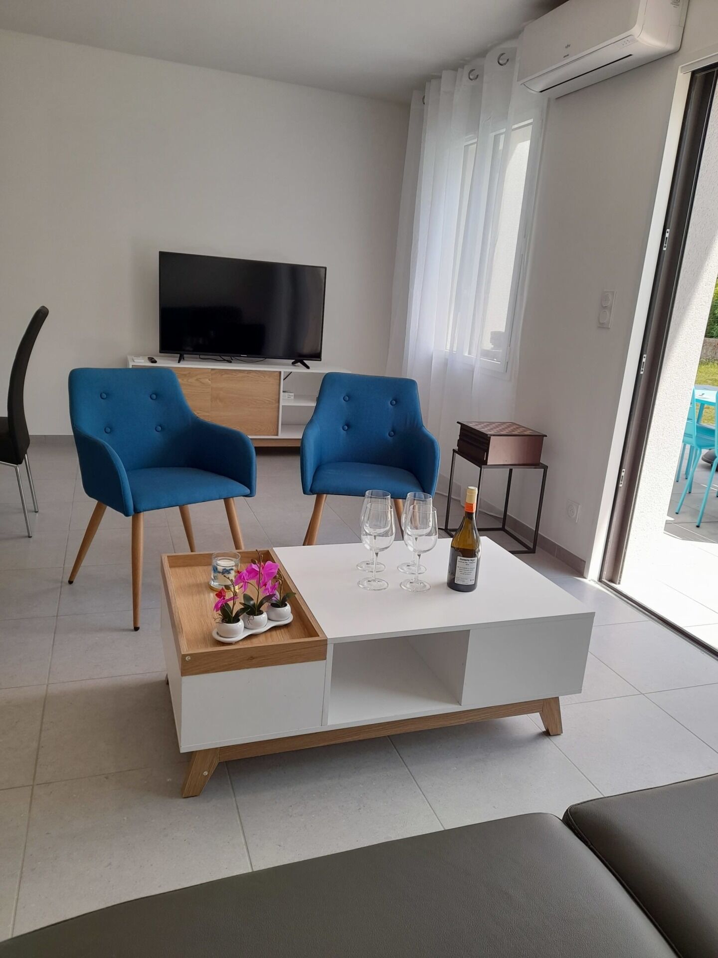 Un salon moderne avec une table basse blanche, deux fauteuils bleus, un téléviseur à écran plat et une porte-fenêtre coulissante donnant sur un espace extérieur.