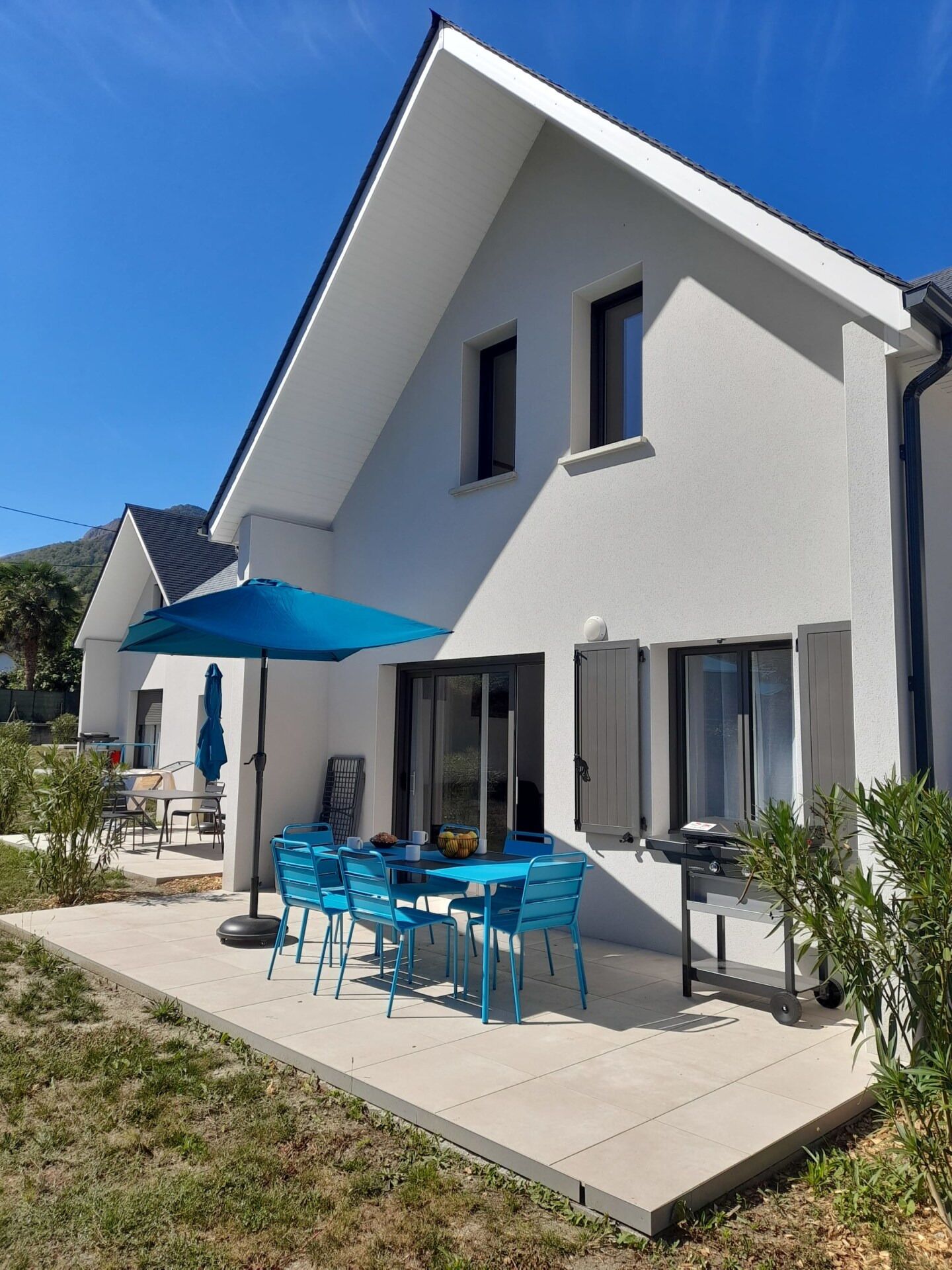 Une maison moderne à deux étages dotée d’un patio comprenant une table et des chaises, un barbecue et un parasol bleu.