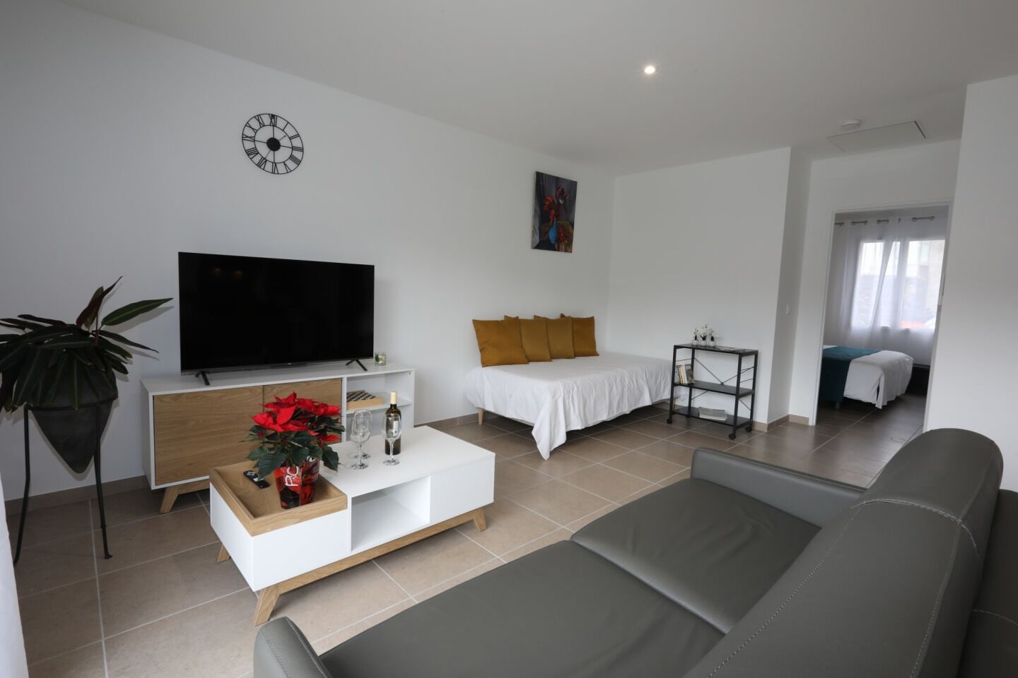 A modern living room with a flat-screen TV, a sofa, a coffee table with a plant, and a bed with a white bedspread and orange pillows.