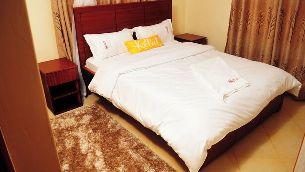 A neatly made bed with white linens and a yellow pillow, a wooden nightstand, and a brown rug. - Double N Suites Kakamega (Kakamega)