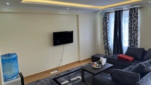 Uma sala de estar com televisão de tela plana fixada na parede, um sofá cinza com um travesseiro vermelho, uma mesa de centro e um dispensador de água.