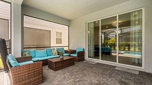 A covered patio area with wicker furniture, a glass door leading to a pool, and a view of greenery.
