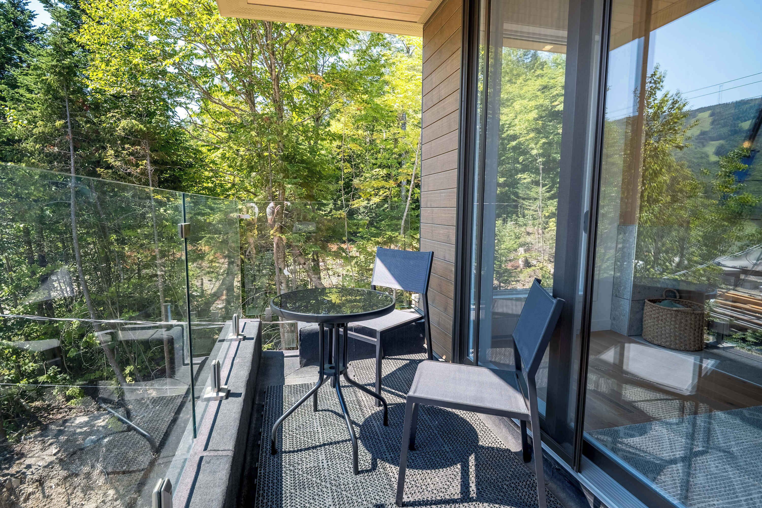 Ein Balkon mit Glasgeländer, einem runden Tisch, zwei Stühlen und Blick auf ein bewaldetes Gebiet.