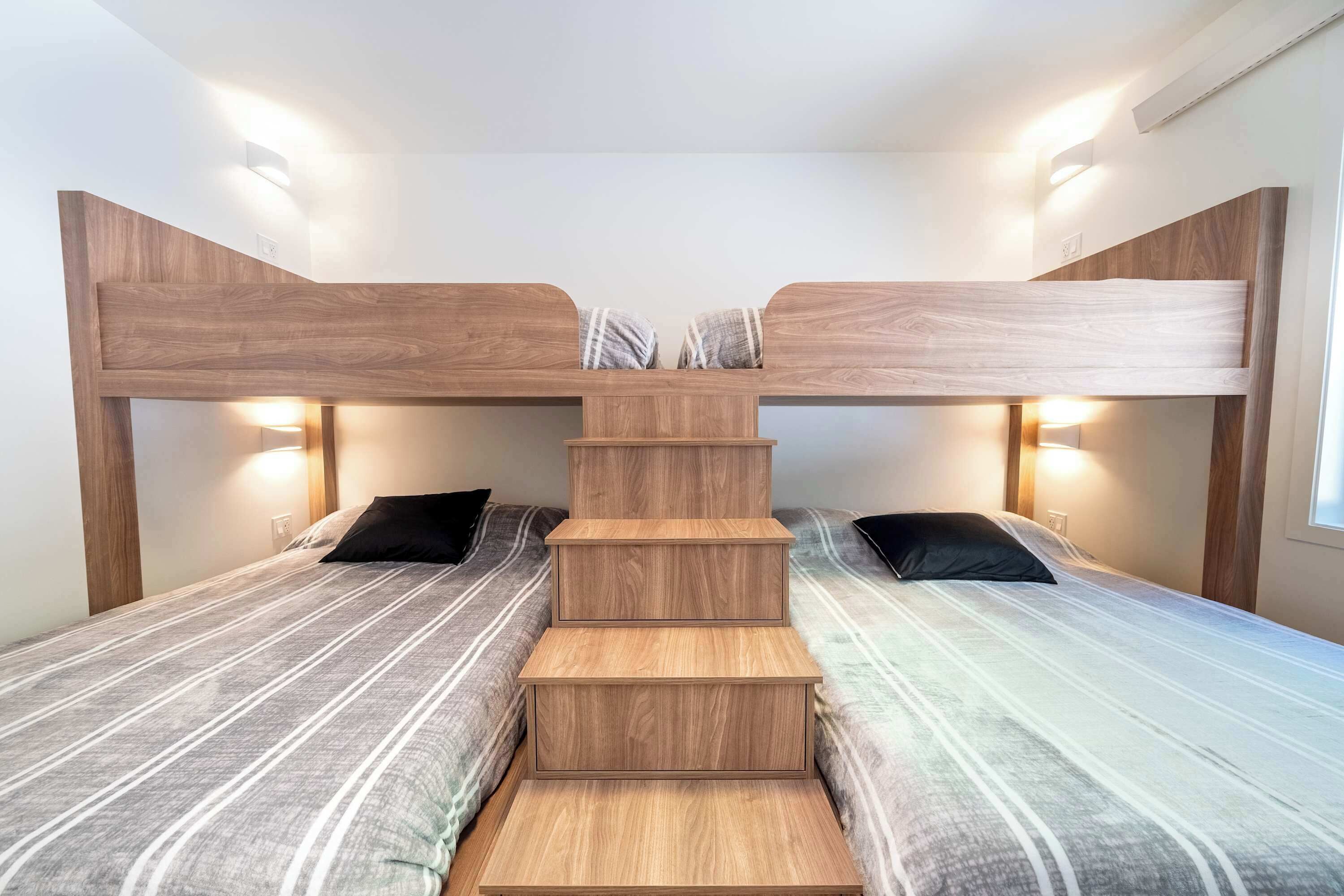 A room with two bunk beds, each with a wooden headboard and a light-colored striped bedspread. There is a wooden staircase connecting the beds.