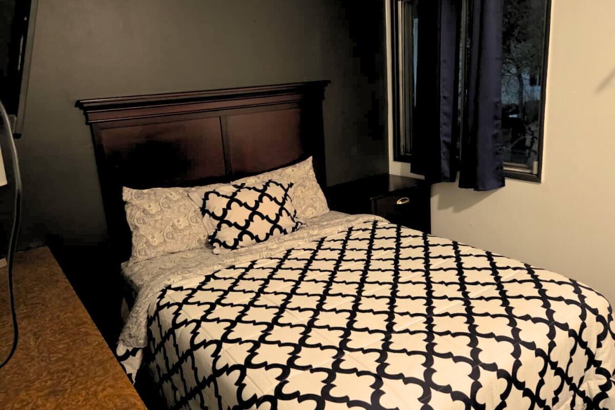 A bedroom with a dark wooden headboard, a patterned bedspread, and a window with dark curtains.
