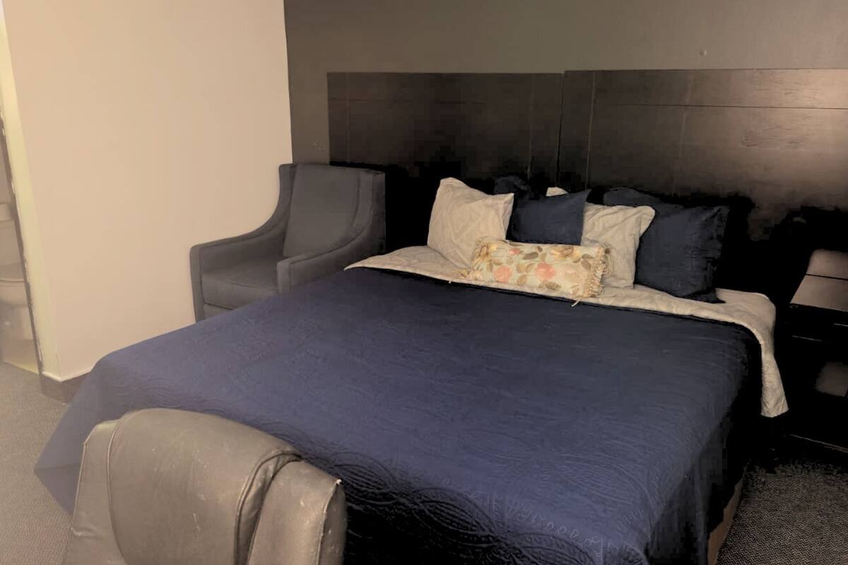 A hotel room with a dark blue bedspread, a grey armchair, and a headboard.
