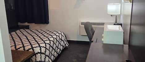 Habitación de hotel pequeña con cama, escritorio, silla, lámpara y radiador.