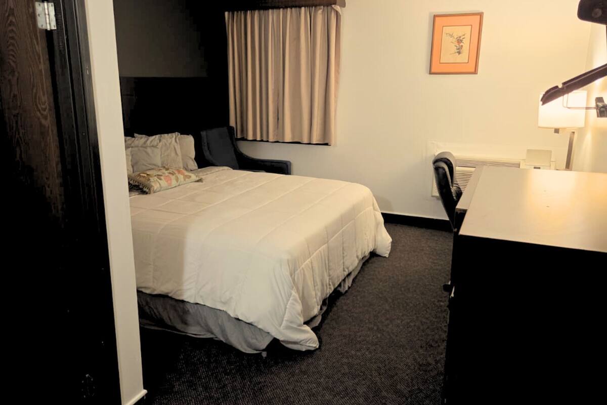 A hotel room with a bed, a desk, a chair, a radiator, and a curtain.