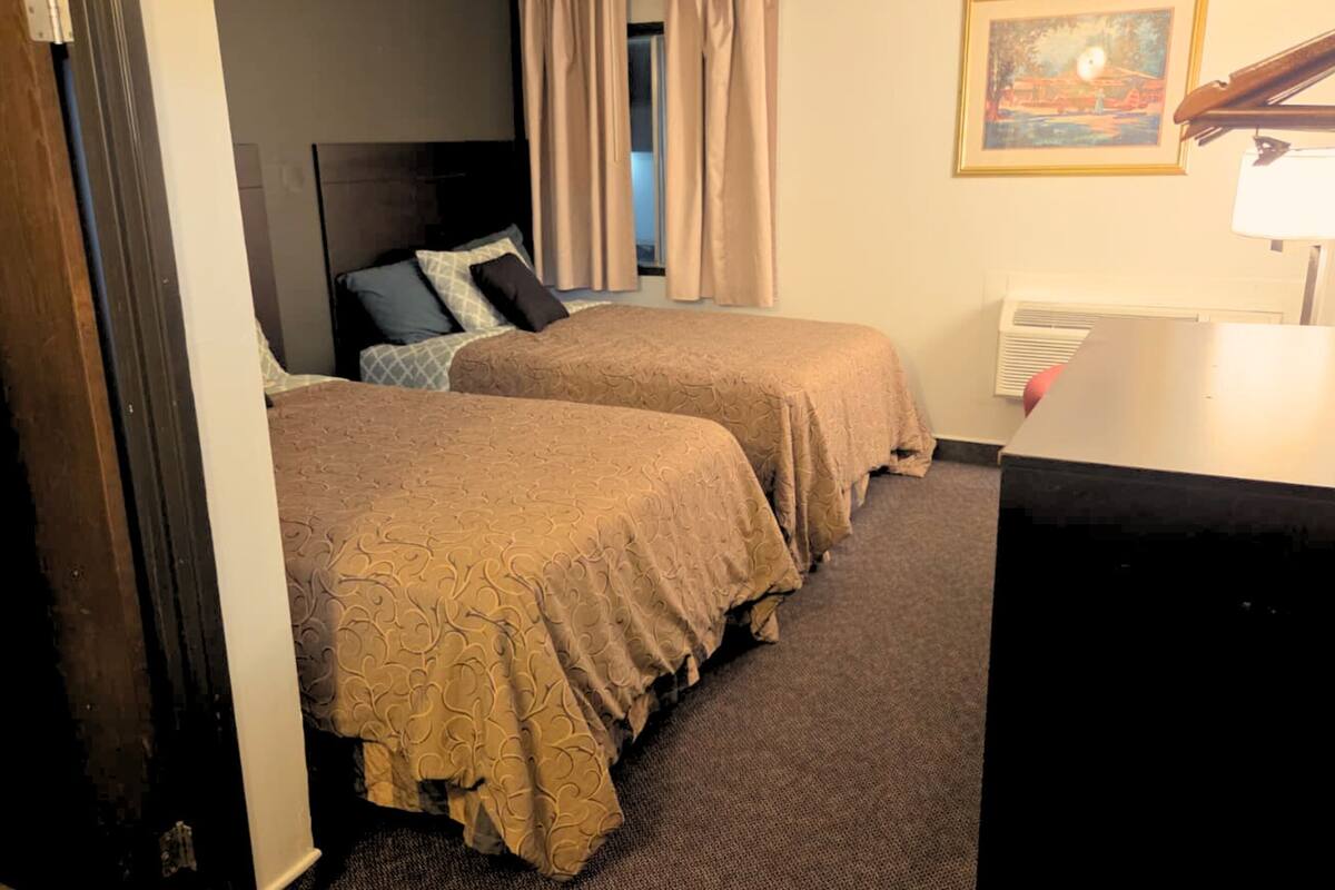 A hotel room with two beds, a desk, a lamp, a painting on the wall, and a window with curtains.