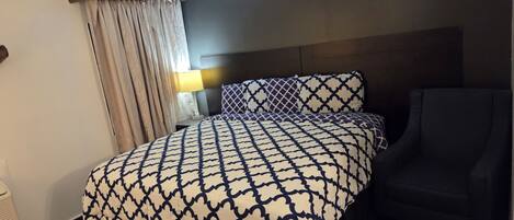 A hotel room with a bed, a chair, a nightstand, a lamp, and a window with curtains.