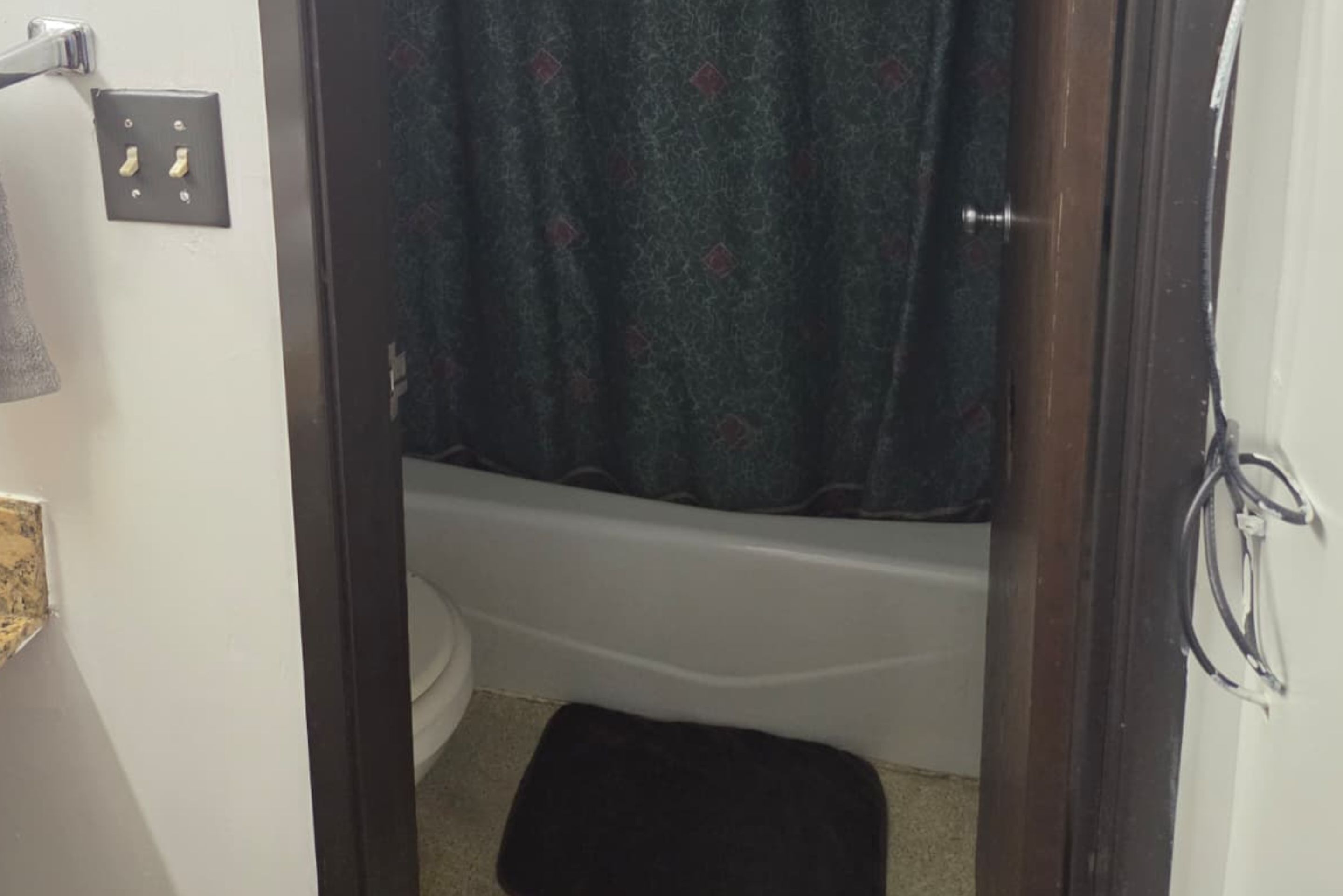 A bathroom with a white bathtub, a black towel on the floor, and a shower curtain with a pattern.