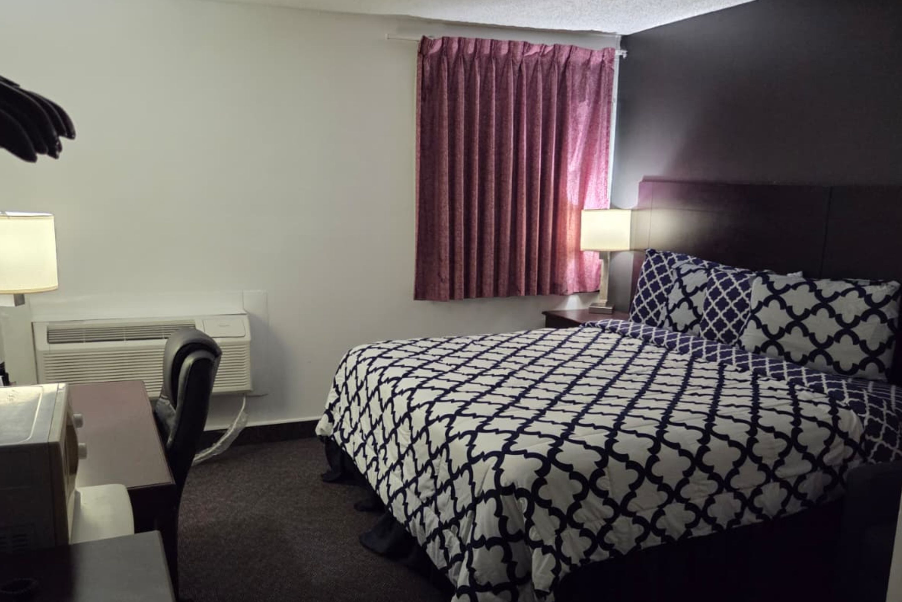 A hotel room with a bed, a desk, a chair, a lamp, and a window with curtains.