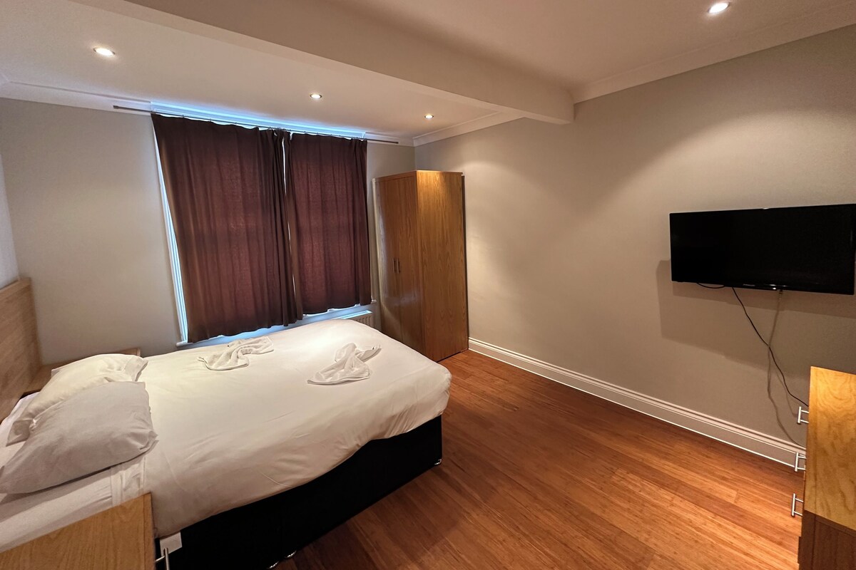 Ein Hotelzimmer mit einem Bett, einem Kleiderschrank, einem an der Wand montierten Fernseher und einem Fenster mit Vorhängen.