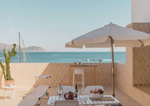A terrace with a table set for two, a large umbrella, and a view of the sea. - Moll Petit Apartments (Can Picafort)