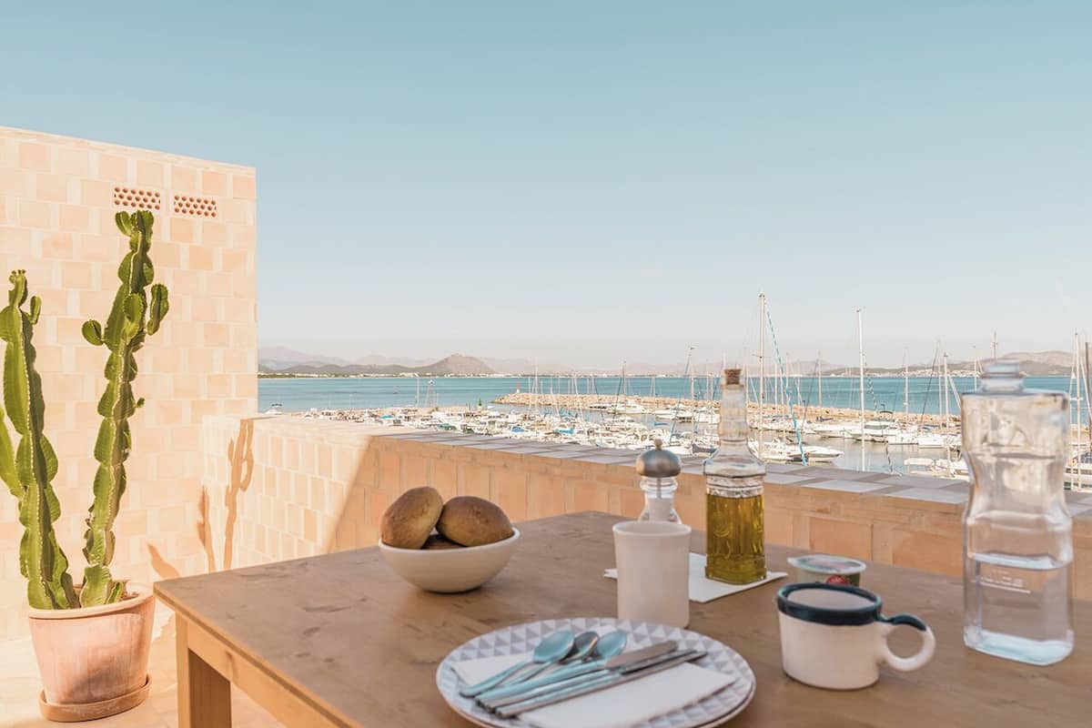 Mesa dispuesta con pan, un bol, una jarrón, una taza y una botella de agua, con vista a un puerto deportivo con barcos.