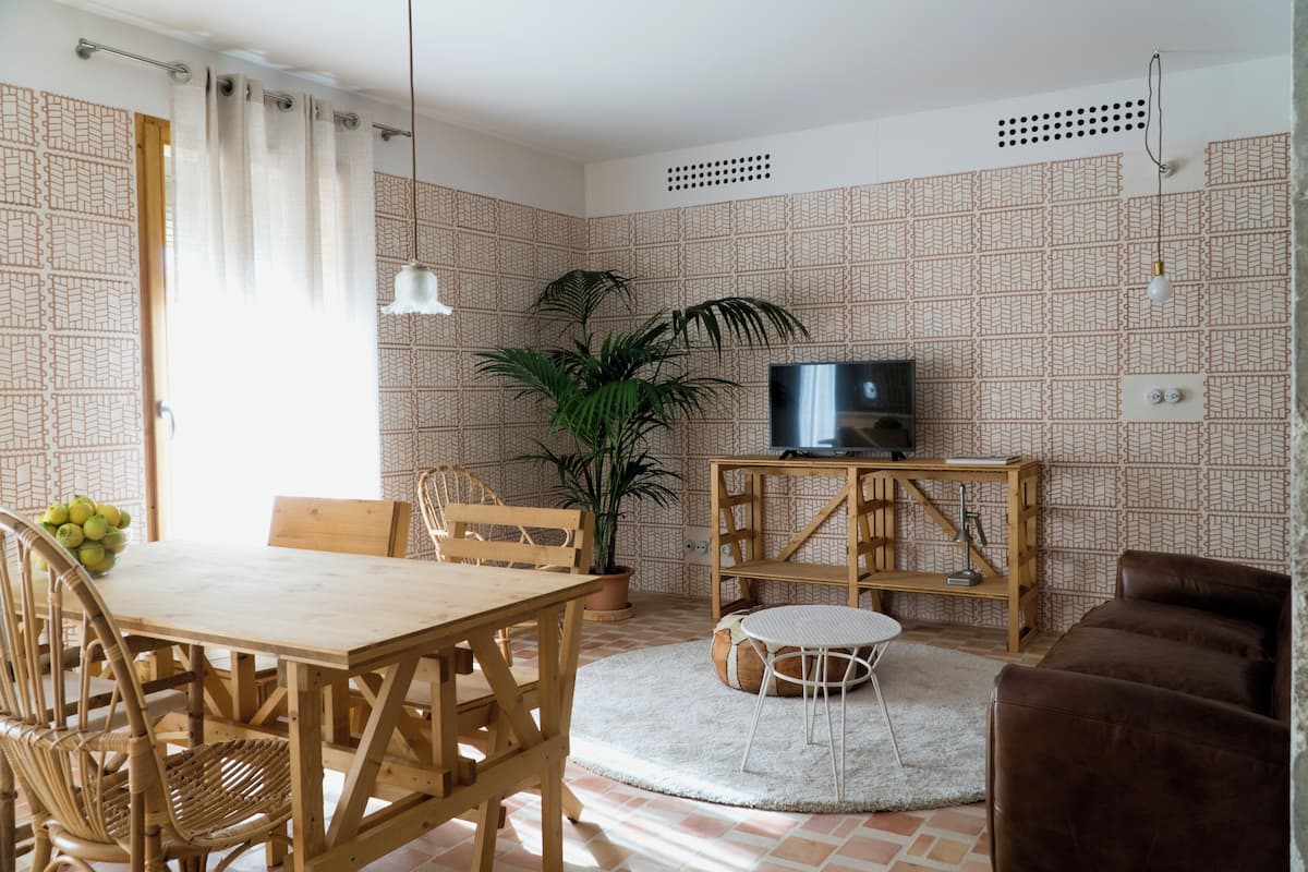 Una sala de estar con una mesa de comedor de madera, sillas, un televisor sobre un mueble de madera, una planta en maceta y un papel tapiz estampado.