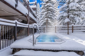 A snow-covered outdoor hot tub area with a wooden fence and snow-laden trees in the background.