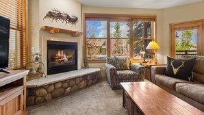 A cozy living room with a fireplace, comfortable seating, and large windows offering a view of the outdoors.