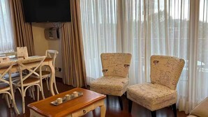 Una sala de estar con un sofá, dos sillones, una mesa de centro de madera, un televisor montado en la pared y un comedor con mesa y sillas.