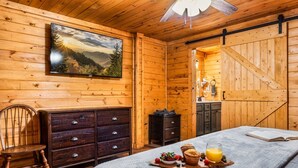 A cozy wooden cabin room with a bed, a dresser, a chair, a breakfast tray with fruits and juice, and a painting of a mountain landscape. - 8 Miles to Pigeon Forge, 15 Miles to Gatlinburg, Sleeps 8, Pool Table, Hot Tub, Game Room (Sevierville)