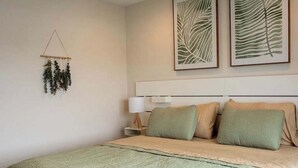 Un lit bien fait, recouvert d’une couvre-lit verte et agrémenté de deux oreillers, une table de chevet avec une lampe, et deux motifs de feuilles encadrés accrochés au mur.
