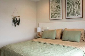 Un lit bien fait, recouvert d’une couvre-lit verte et agrémenté de deux oreillers, une table de chevet avec une lampe, et deux motifs de feuilles encadrés accrochés au mur.