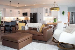 A modern kitchen with white cabinets, a black refrigerator, and pendant lighting. There is a brown sectional sofa with a white throw blanket and a white rocking chair.