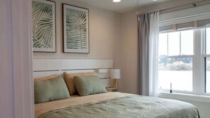 Une chambre à coucher avec un lit, deux oreillers, une table de chevet avec une lampe et des œuvres d’art encadrées accrochées au mur.