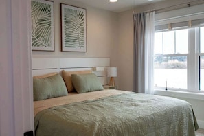 Een slaapkamer met een bed, twee kussens, een nachtkastje met een lamp en een kunstwerk in een lijst aan de muur.