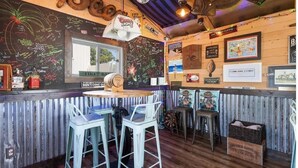 Un bar au décor rustique, avec un intérieur en bois, des tabourets de bar, un mur noir où sont écrits des messages à la craie et une enseigne Coors Light.
