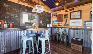 Un bar au décor rustique, avec un intérieur en bois, des tabourets de bar, un mur noir où sont écrits des messages à la craie et une enseigne Coors Light.