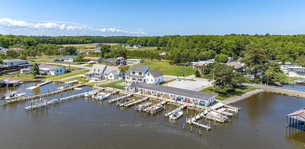 Un port de plaisance avec des bateaux amarrés, un bâtiment blanc doté d’un grand garage, et un quartier résidentiel avec des maisons et de la verdure en arrière-plan.
