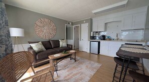 A living room with a sofa, coffee table, and a dining area with a table and chairs. There is a kitchen area with cabinets and appliances.