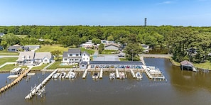 A marina with docked boats, a large house with a covered porch, and a forested hill in the background.