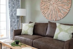 A brown leather sofa with patterned cushions, a round wall decoration, and a side table with a plant and a lamp.
