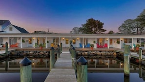 A waterfront hotel with a wooden dock leading to a stone-lined waterway, featuring outdoor seating and a covered porch area.