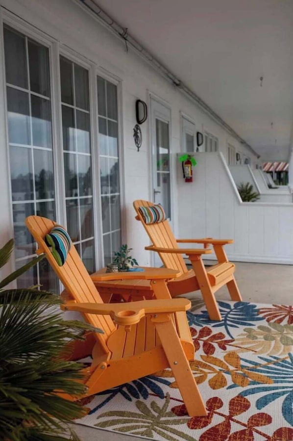 A covered porch with two wooden Adirondack chairs and a small table, a patterned rug, and a potted plant.